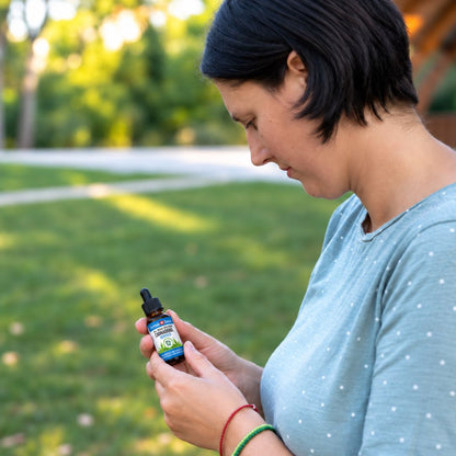 Frau im Park liest das Etikett einer kleinen Flasche CBD-Öl, in hellblauem Punkte-T-Shirt mit roten und grünen Armbändern